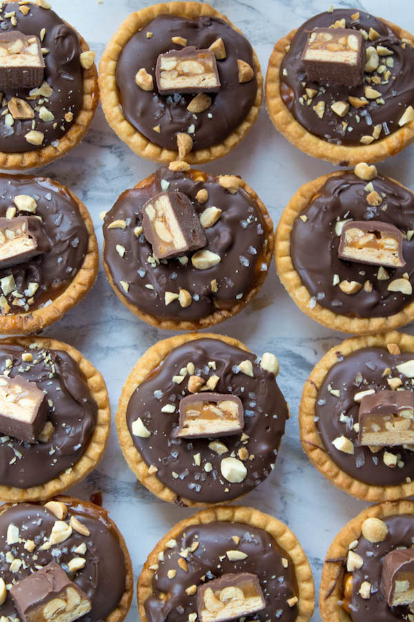 Mini Salted Caramel Snickers Tarts! Mini pre-baked tart shells with a salted caramel peanut filling and a chocolate peanut butter topping. Finish it off with chopped snickers and more peanuts, and you have the easiest/most decadent and delicious, bite-sized dessert!