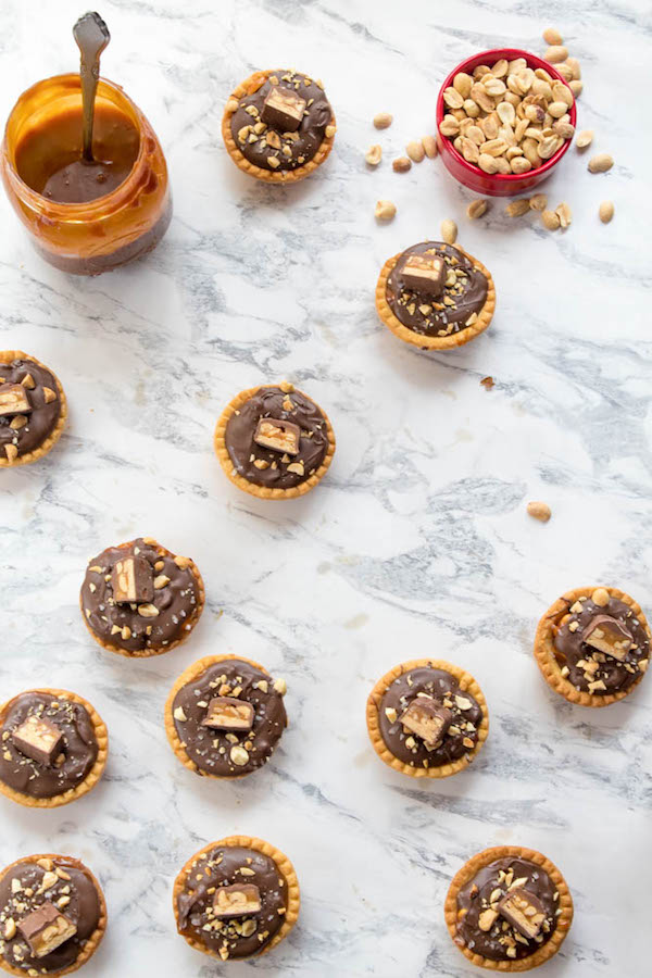 Mini Salted Caramel Snickers Tarts! Mini pre-baked tart shells with a salted caramel peanut filling and a chocolate peanut butter topping. Finish it off with chopped snickers and more peanuts, and you have the easiest/most decadent and delicious, bite-sized dessert!