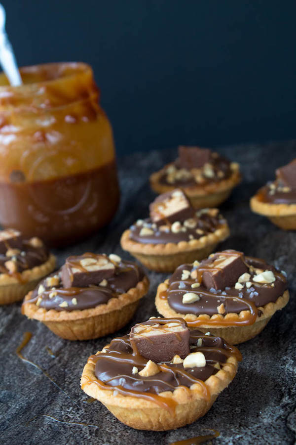 Mini Salted Caramel Snickers Tarts! Mini pre-baked tart shells with a salted caramel peanut filling and a chocolate peanut butter topping. Finish it off with chopped snickers and more peanuts, and you have the easiest/most decadent and delicious, bite-sized dessert!