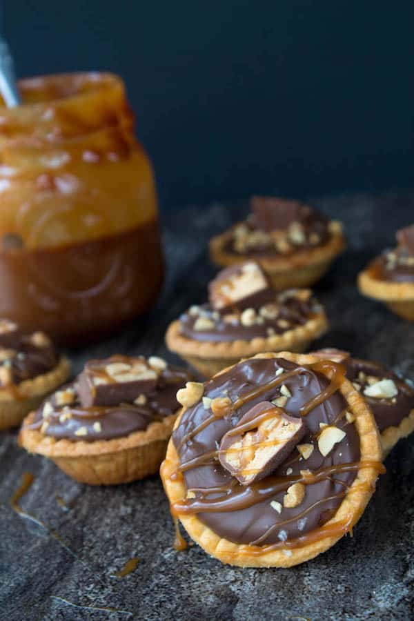 Mini Salted Caramel Snickers Tarts! Mini pre-baked tart shells with a salted caramel peanut filling and a chocolate peanut butter topping. Finish it off with chopped snickers and more peanuts, and you have the easiest/most decadent and delicious, bite-sized dessert!