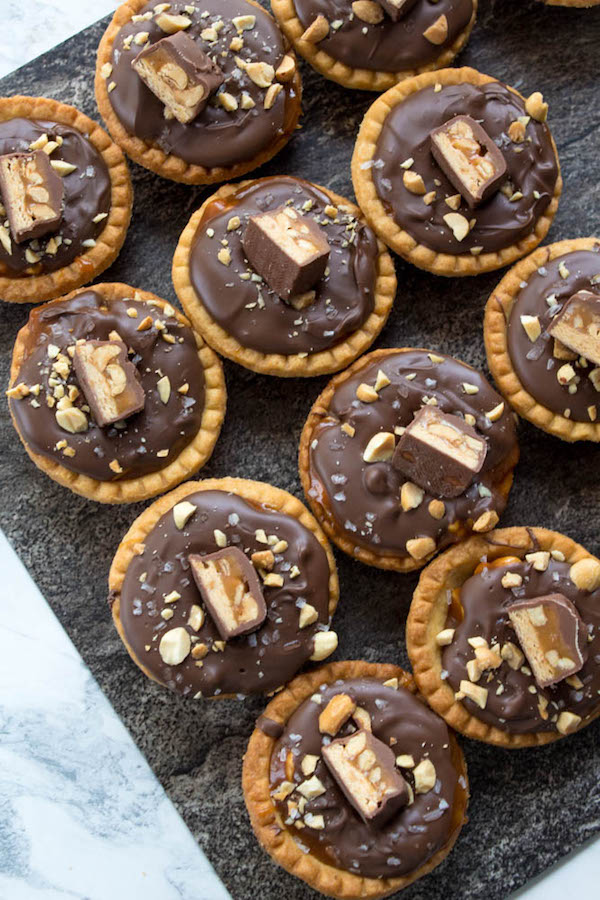 Mini Salted Caramel Snickers Tarts! Mini pre-baked tart shells with a salted caramel peanut filling and a chocolate peanut butter topping. Finish it off with chopped snickers and more peanuts, and you have the easiest/most decadent and delicious, bite-sized dessert!