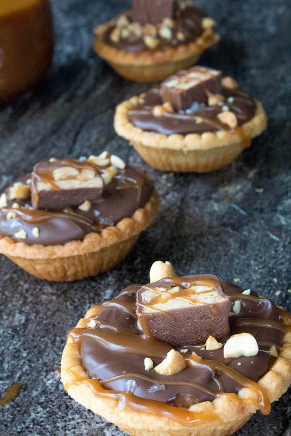 Mini Salted Caramel Snickers Tarts! Mini pre-baked tart shells with a salted caramel peanut filling and a chocolate peanut butter topping. Finish it off with chopped snickers and more peanuts, and you have the easiest/most decadent and delicious, bite-sized dessert!