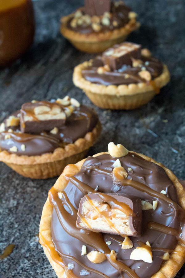 Mini Salted Caramel Snickers Tarts! Mini pre-baked tart shells with a salted caramel peanut filling and a chocolate peanut butter topping. Finish it off with chopped snickers and more peanuts, and you have the easiest/most decadent and delicious, bite-sized dessert!