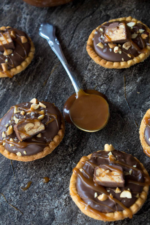 Mini Salted Caramel Snickers Tarts! Mini pre-baked tart shells with a salted caramel peanut filling and a chocolate peanut butter topping. Finish it off with chopped snickers and more peanuts, and you have the easiest/most decadent and delicious, bite-sized dessert!