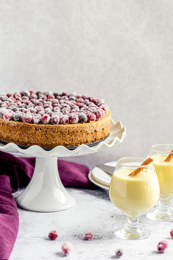 eggnog cheesecake on a cake stand and two glasses of eggnog