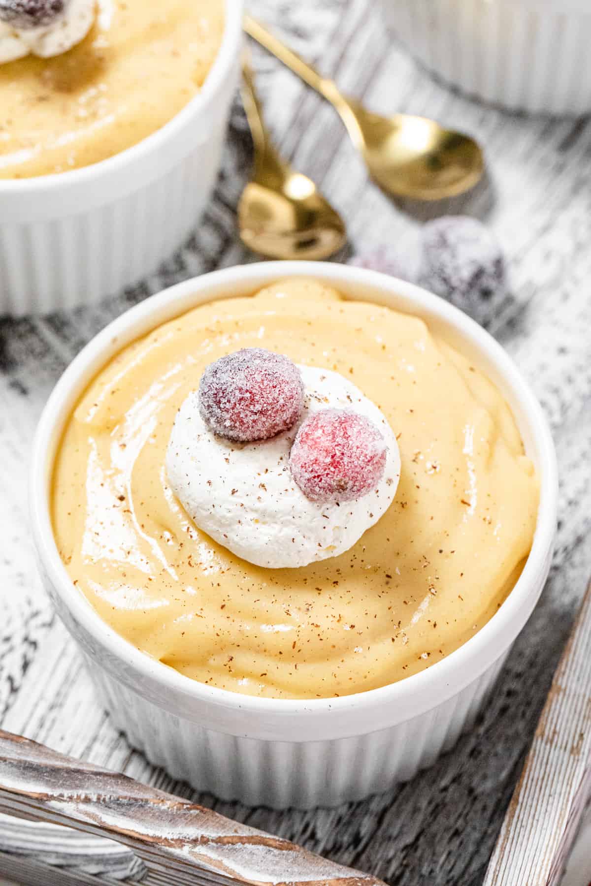 close up of pudding topped with whipped cream and sugared cranberries with gold spoons in the background