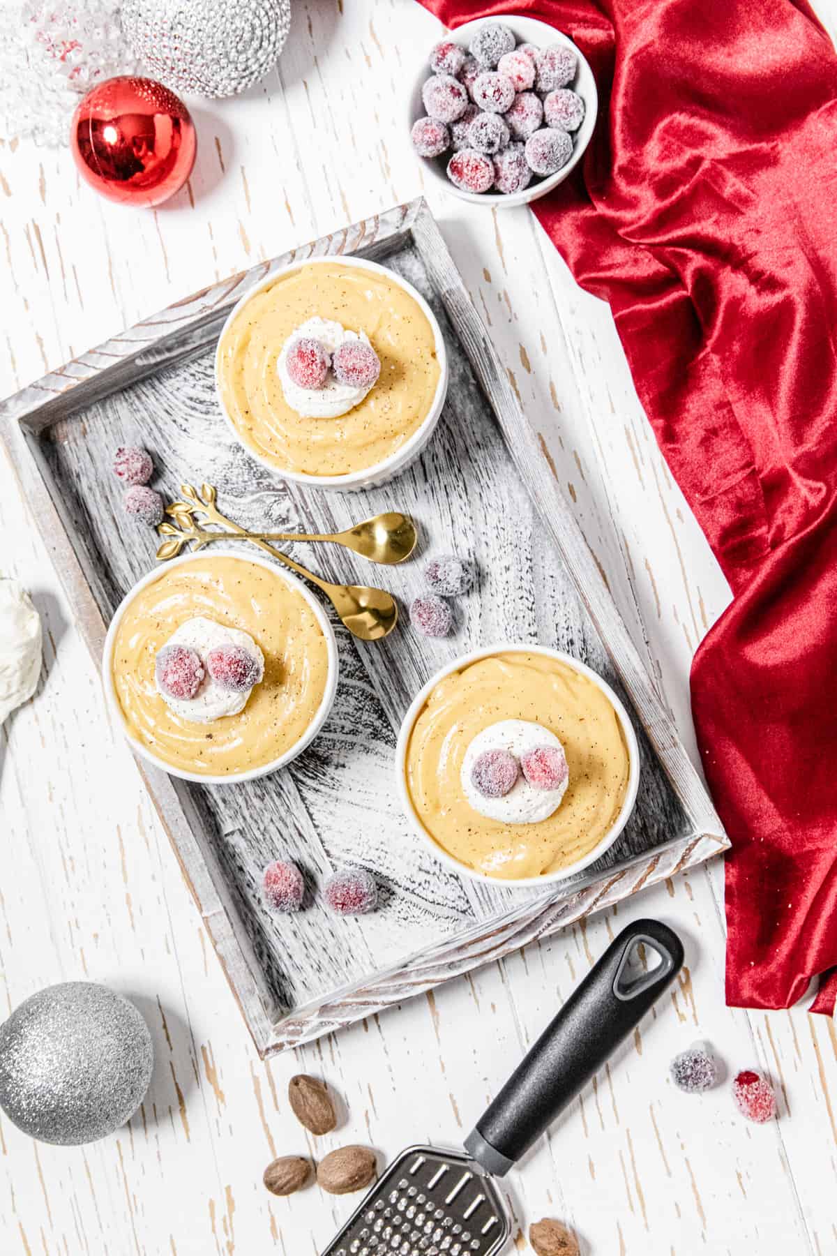 flat-lay of three ramekins with pudding on a wood tray with sugared cranberries, whole nutmegs and red and white ornaments around it