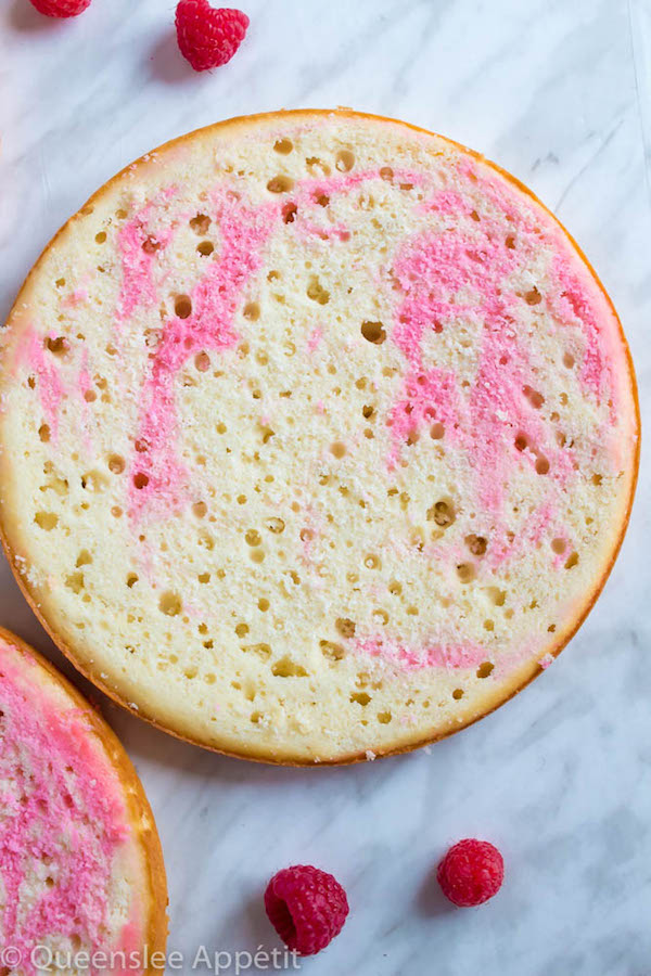 This Vanilla Raspberry Cake has moist and fluffy layers of vanilla cake. Between those layers is a vanilla buttercream with a raspberry sauce. Decorated with a raspberry and vanilla buttercream watercolour design, raspberry buttercream border, garnished with fresh raspberries and more raspberry sauce in the middle.