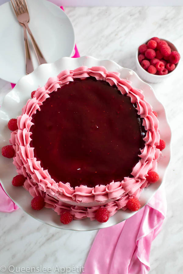 This Vanilla Raspberry Cake has moist and fluffy layers of vanilla cake. Between those layers is a vanilla buttercream with a raspberry sauce. Decorated with a raspberry and vanilla buttercream watercolour design, raspberry buttercream border, garnished with fresh raspberries and more raspberry sauce in the middle.