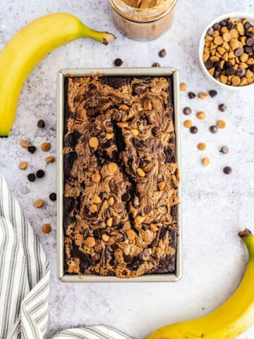 loaf of chocolate and peanut butter swirl banana bread surrounded by two bananas, a jar of peanut butter and a mix of chocolate and peanut butter chips