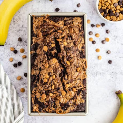 loaf of chocolate and peanut butter swirl banana bread surrounded by two bananas, a jar of peanut butter and a mix of chocolate and peanut butter chips