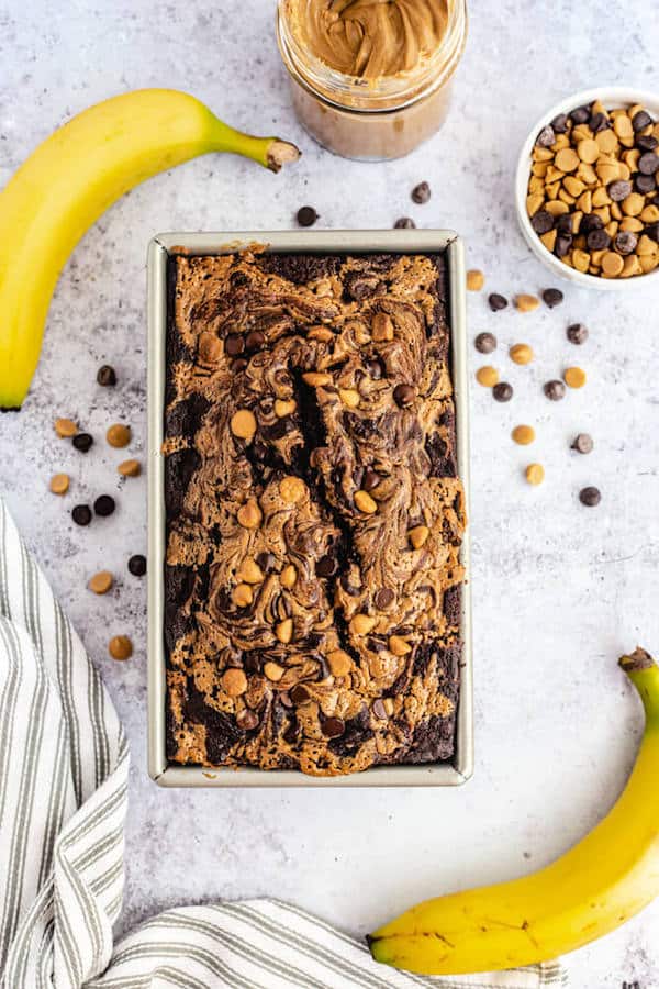 loaf of chocolate and peanut butter swirl banana bread surrounded by two bananas, a jar of peanut butter and a mix of chocolate and peanut butter chips 