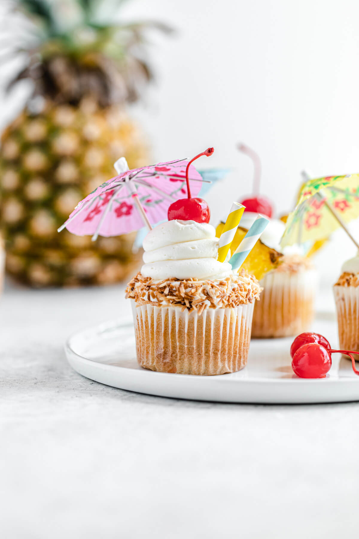 cupcake topped with umbrellas on a white plate with pineapple behind them