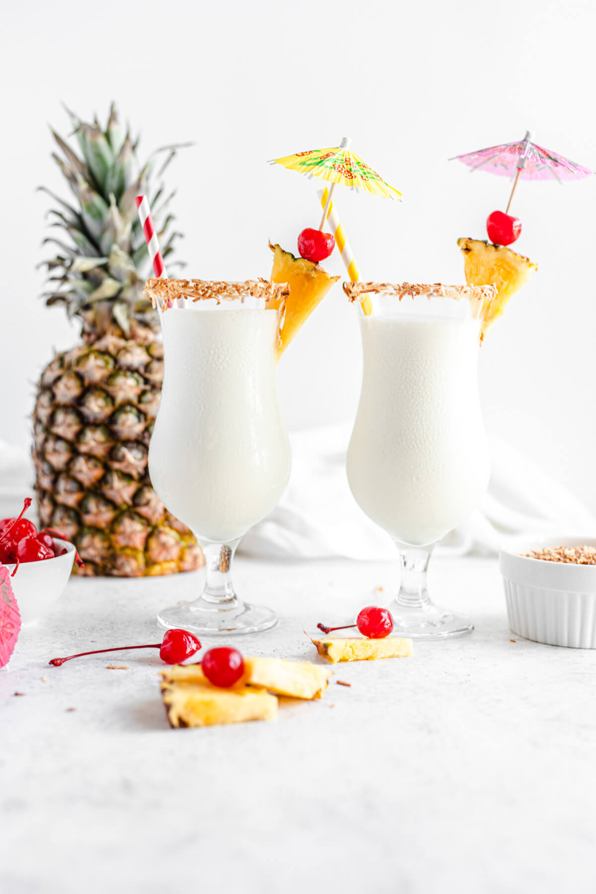 two glasses of milkshake with cherries, toasted coconut and pineapple around