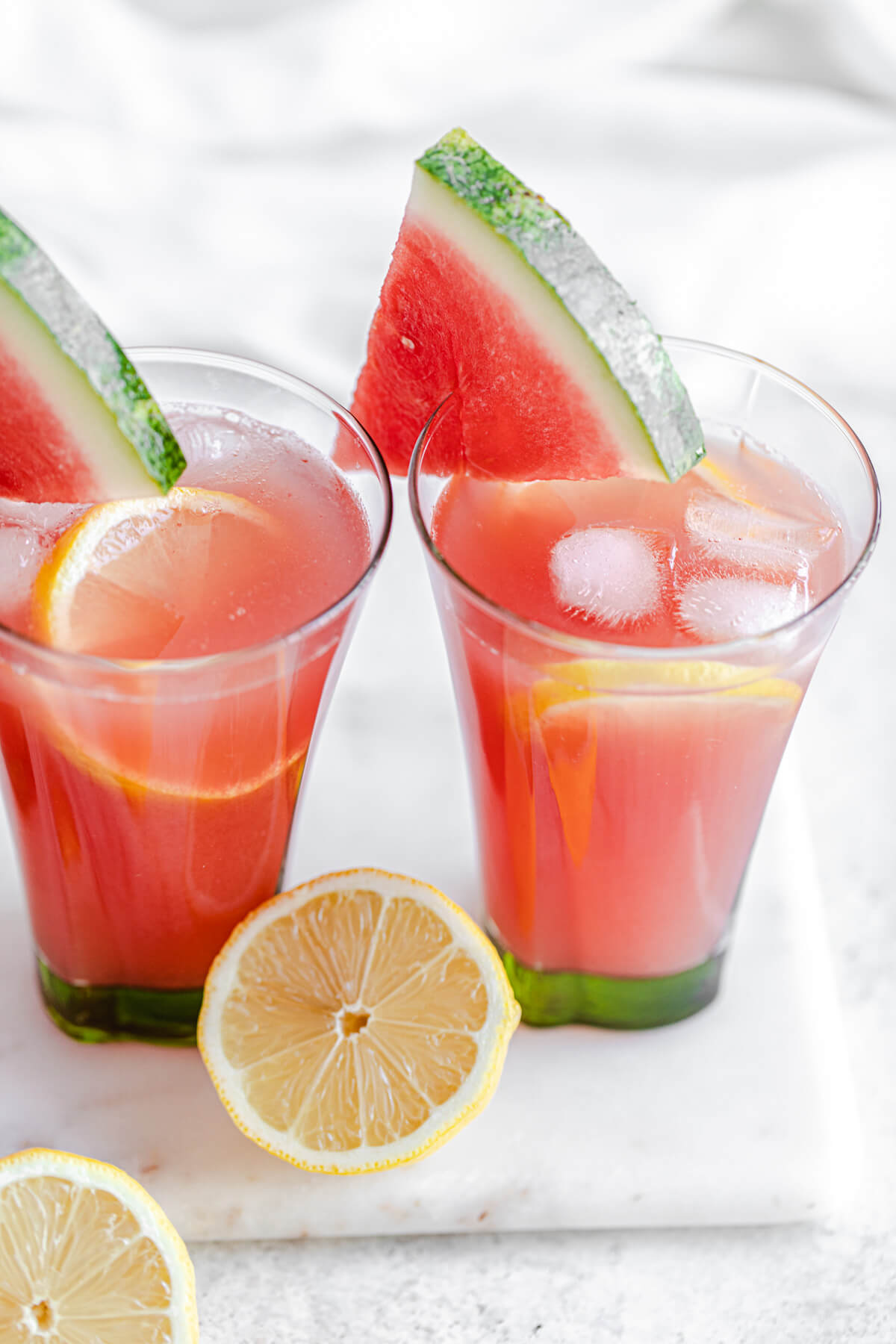 angled view showing lemonade in two glasses with ice, lemon slices and a wedge of watermelon on the rim