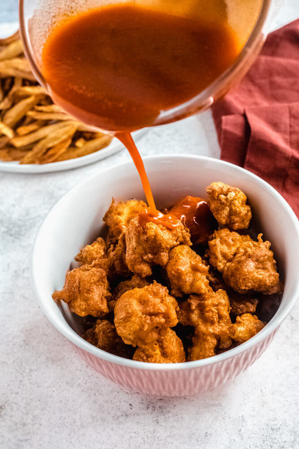 pouring buffalo hot sauce over fried chicken pieces in a bowl