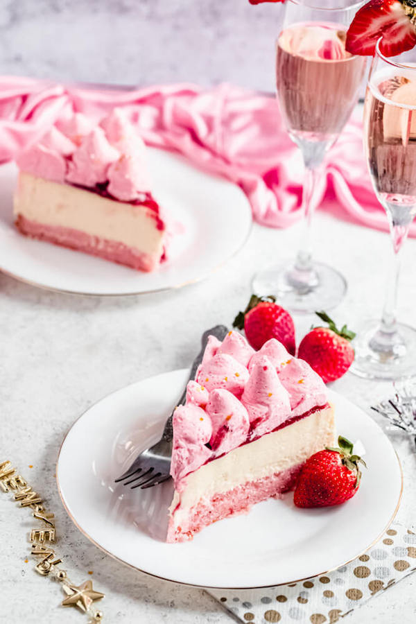 two slices of champagne and strawberry cheesecake on white plates with glasses of pink champagne on the side