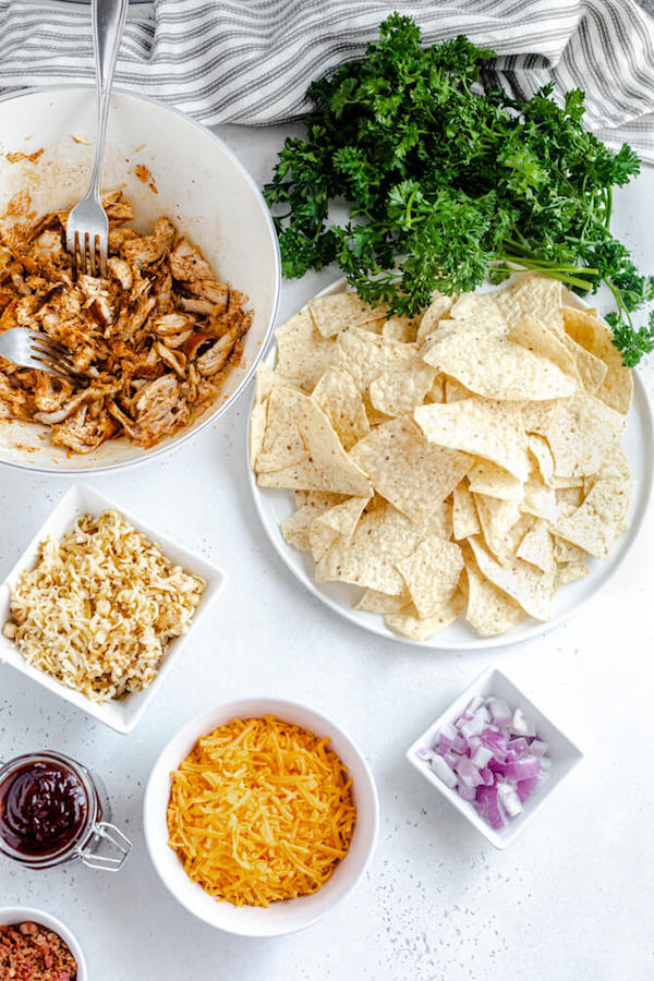 ingredients for nachos - tortilla chips, fresh parsley, diced red onion, crumbled bacon, shredded chicken, shredded cheddar and smoked gouda cheese, honey bbq sauce