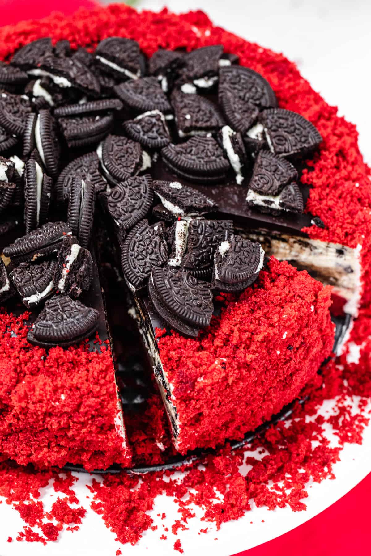 sliced cheesecake covered in red velvet cake crumbs and topped with Oreos on a white cake stand