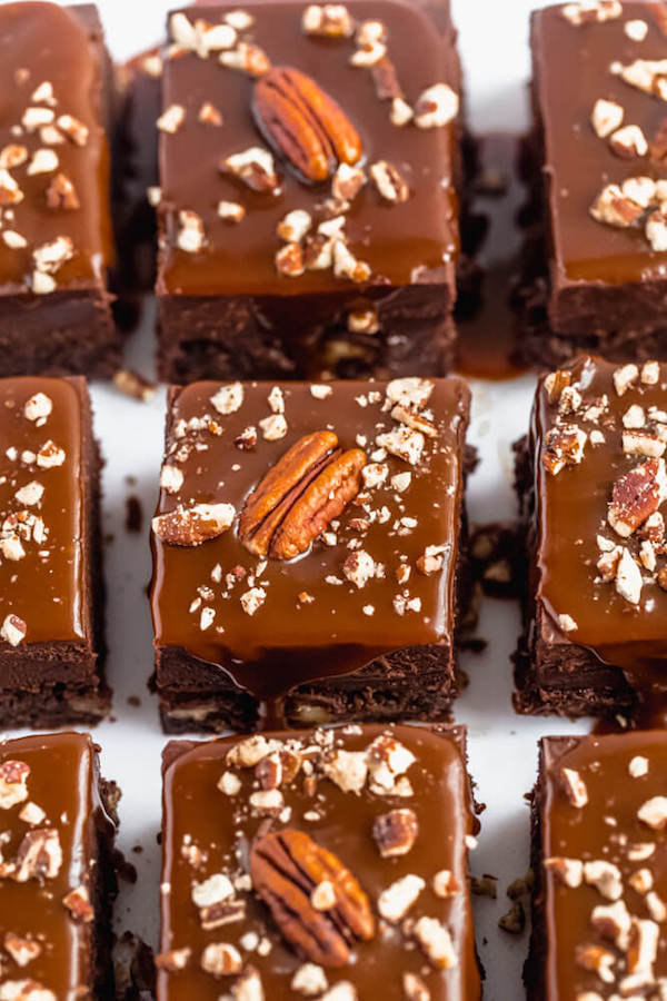 brownies with chocolate chips and chopped pecans baked inside and topped with chocolate fudge, salted caramel sauce and chopped pecans