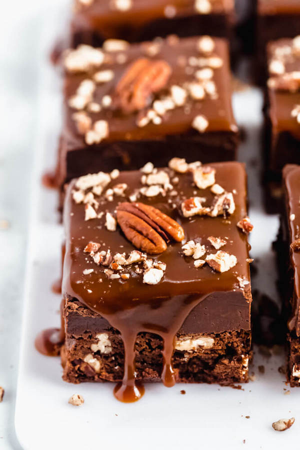 brownies with chocolate chips and chopped pecans baked inside and topped with chocolate fudge, salted caramel sauce and chopped pecans