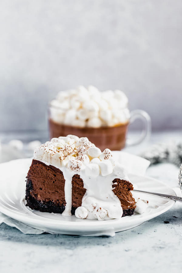 slice of hot chocolate flavoured cheesecake with marshmallow sauce on top and mug of hot chocolate in the back