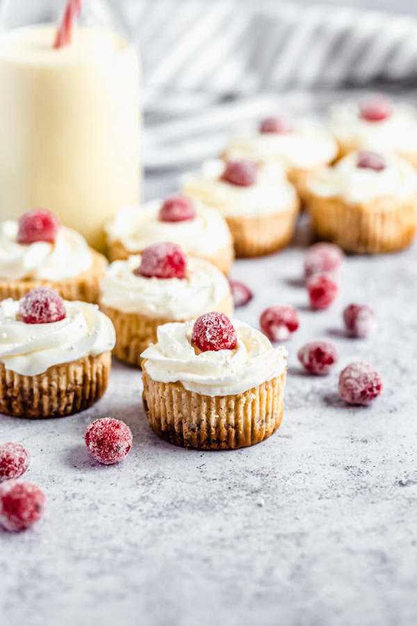 mini eggnog cheesecakes topped with sugared cranberries and a glass of eggnog in the back