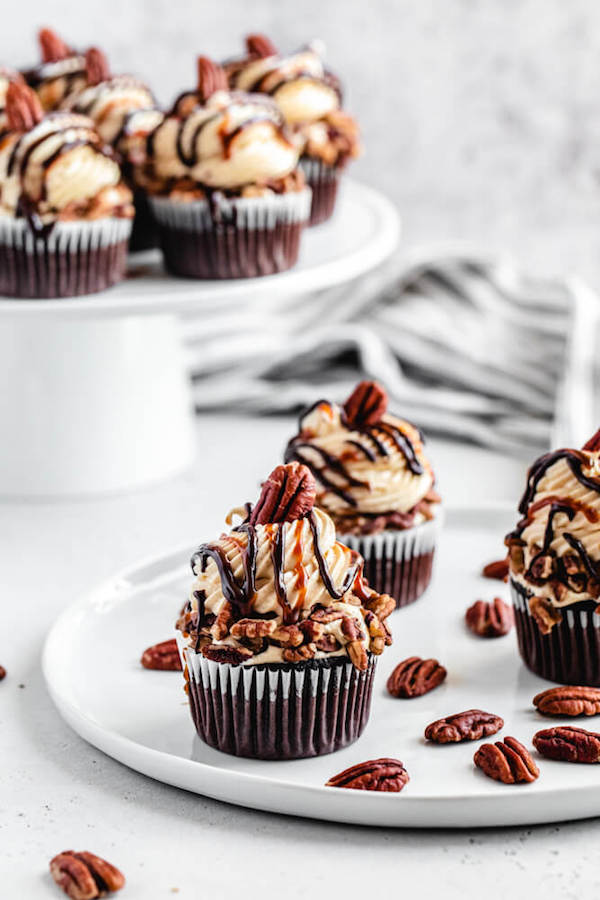 three pecan caramel cupcakes on a white plate with cupcakes on a cake stand in the background 