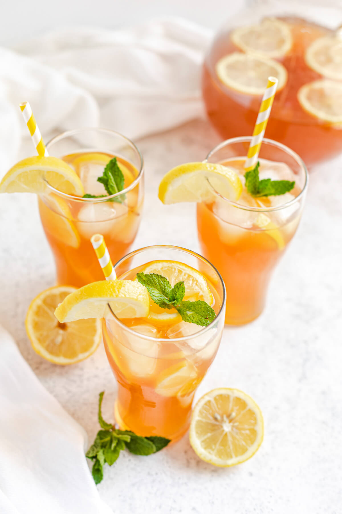 three glasses of lemon flavoured iced tea with striped yellow straws, fresh mint, ice and lemons inside and pitcher in the back