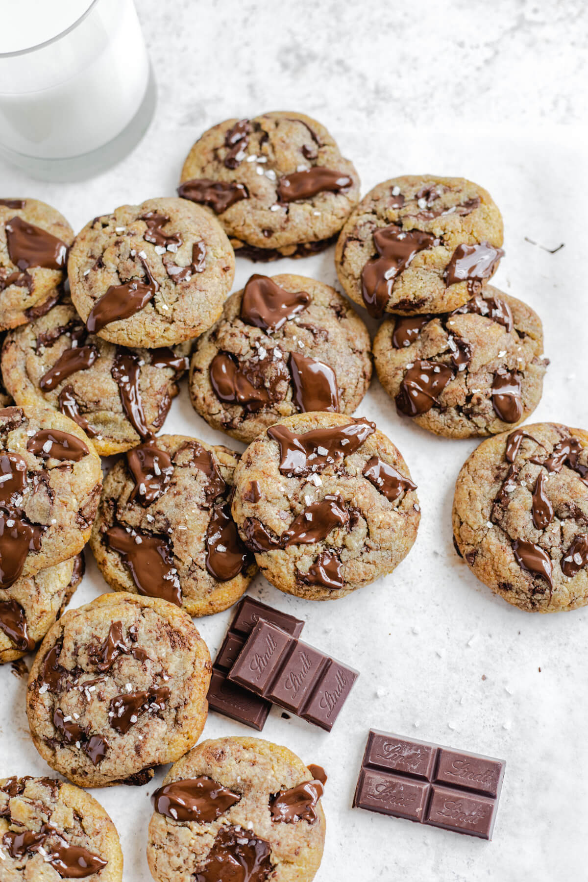 plenty of cookies piled close together with glass of milk in the back and dark chocolate pieces in the foreground