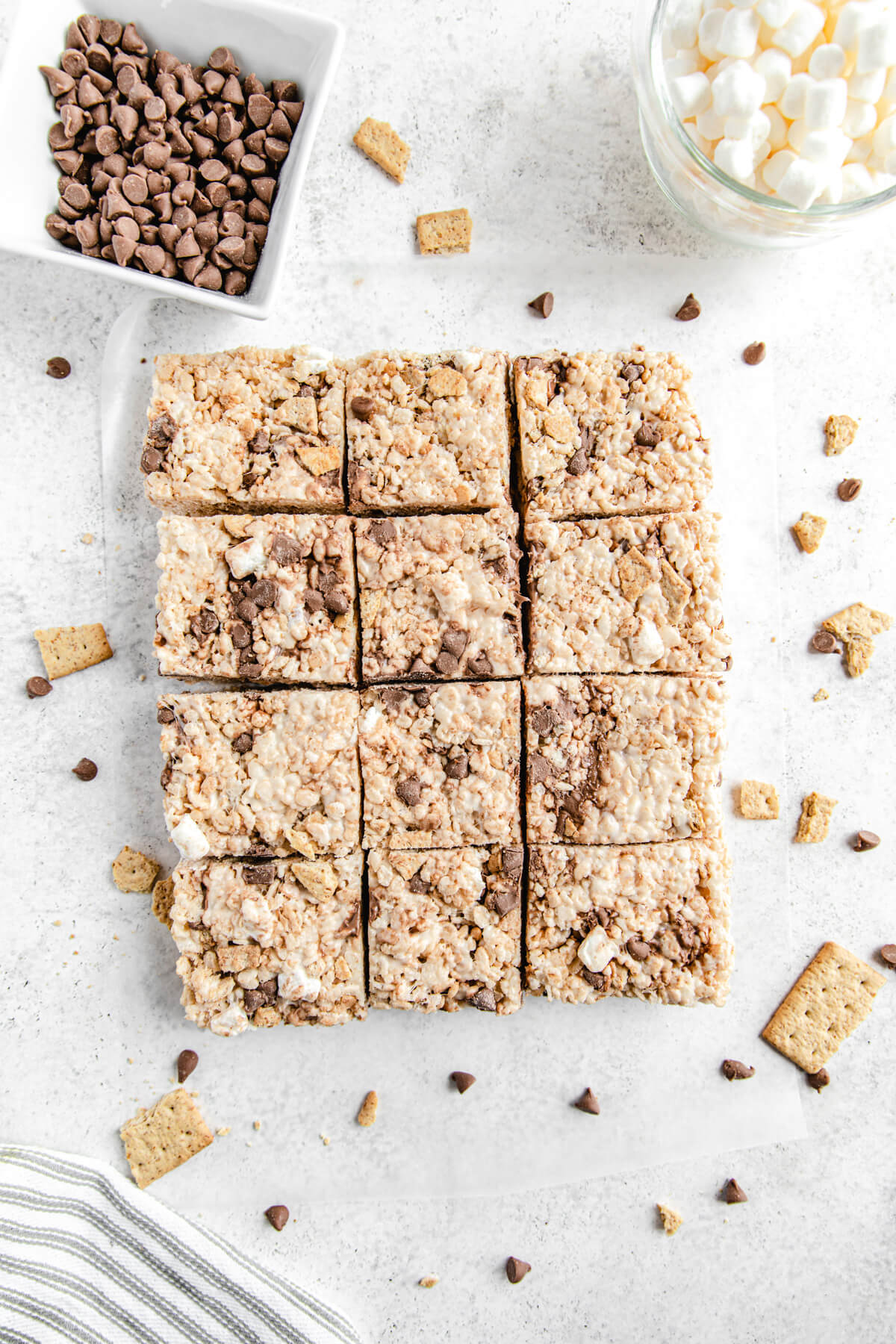 top shot of sliced s'mores flavoured Rice Krispie bars lined up together surrounded by chocolate chips, marshmallows and graham crackers