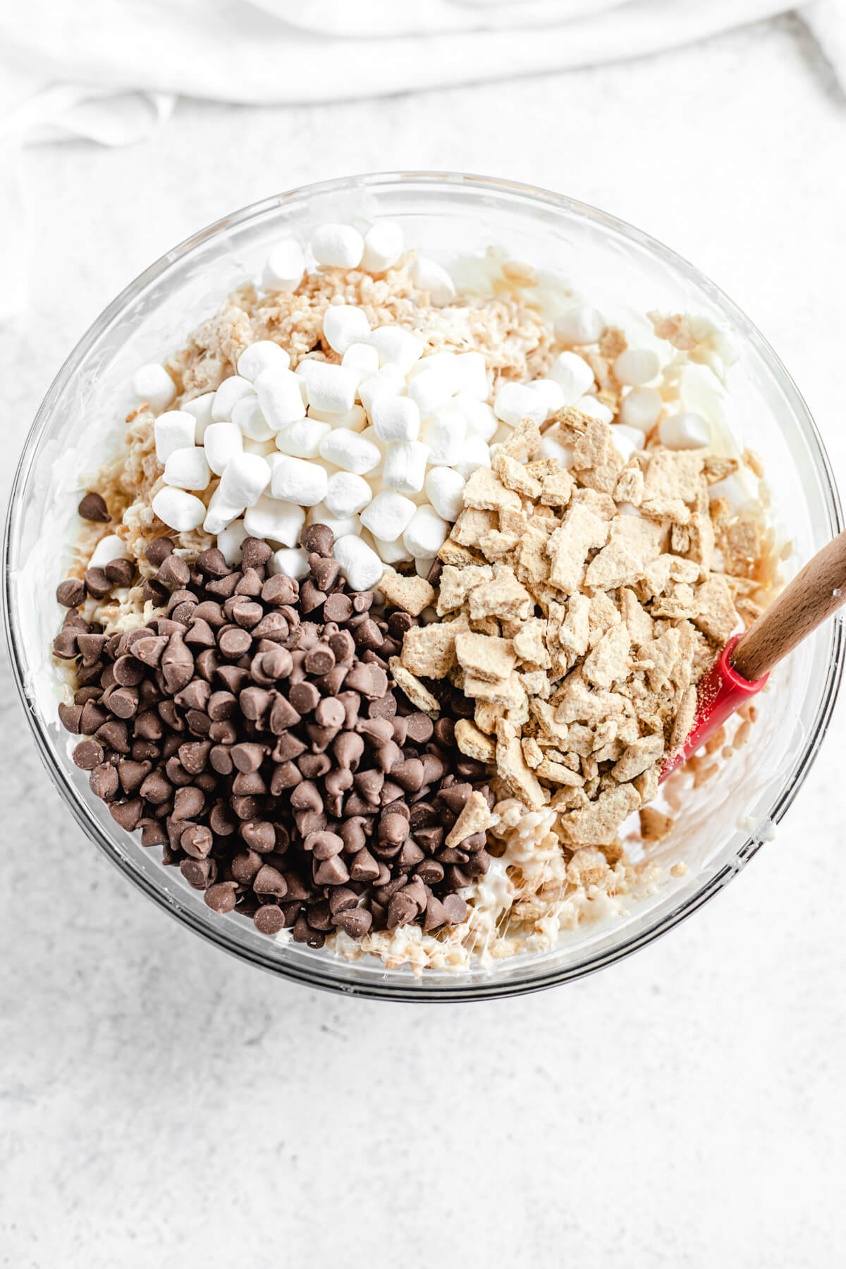 milk chocolate chips, crushed graham crackers and mini marshmallows in a bowl of Rice Krispie treats with rubber spatula inside bowl
