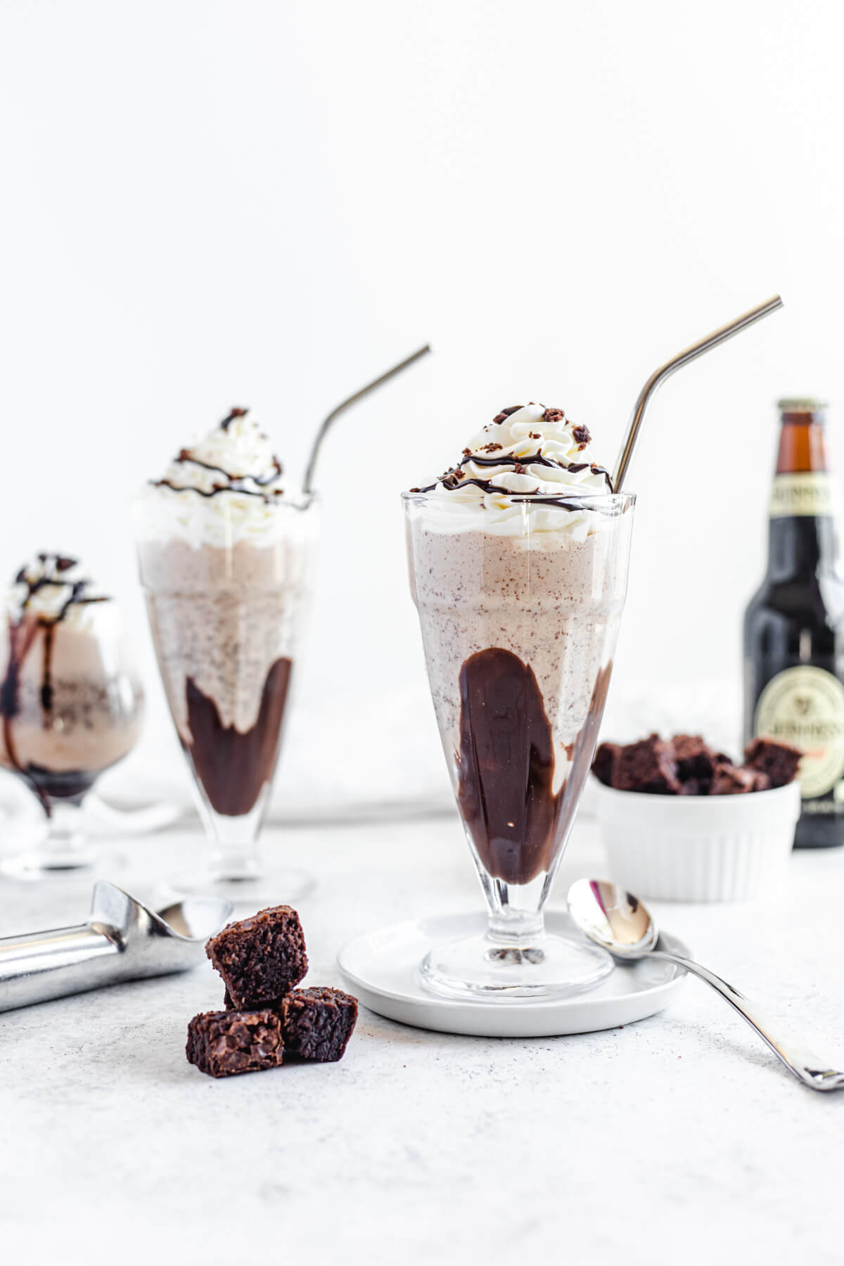 two large glasses of milkshake with a small glass in the background along with brownie chunks and a bottle of beer
