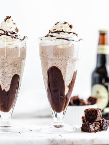 two milkshakes on a rectangle marble board with three brownie chunks beside them and a bottle of beer in the background