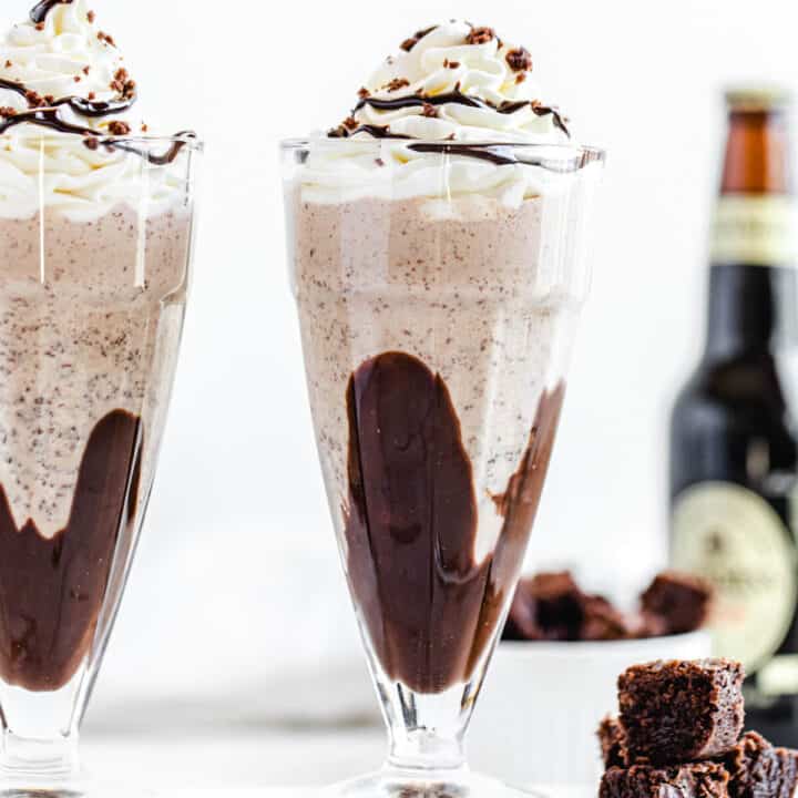 two milkshakes on a rectangle marble board with three brownie chunks beside them and a bottle of beer in the background
