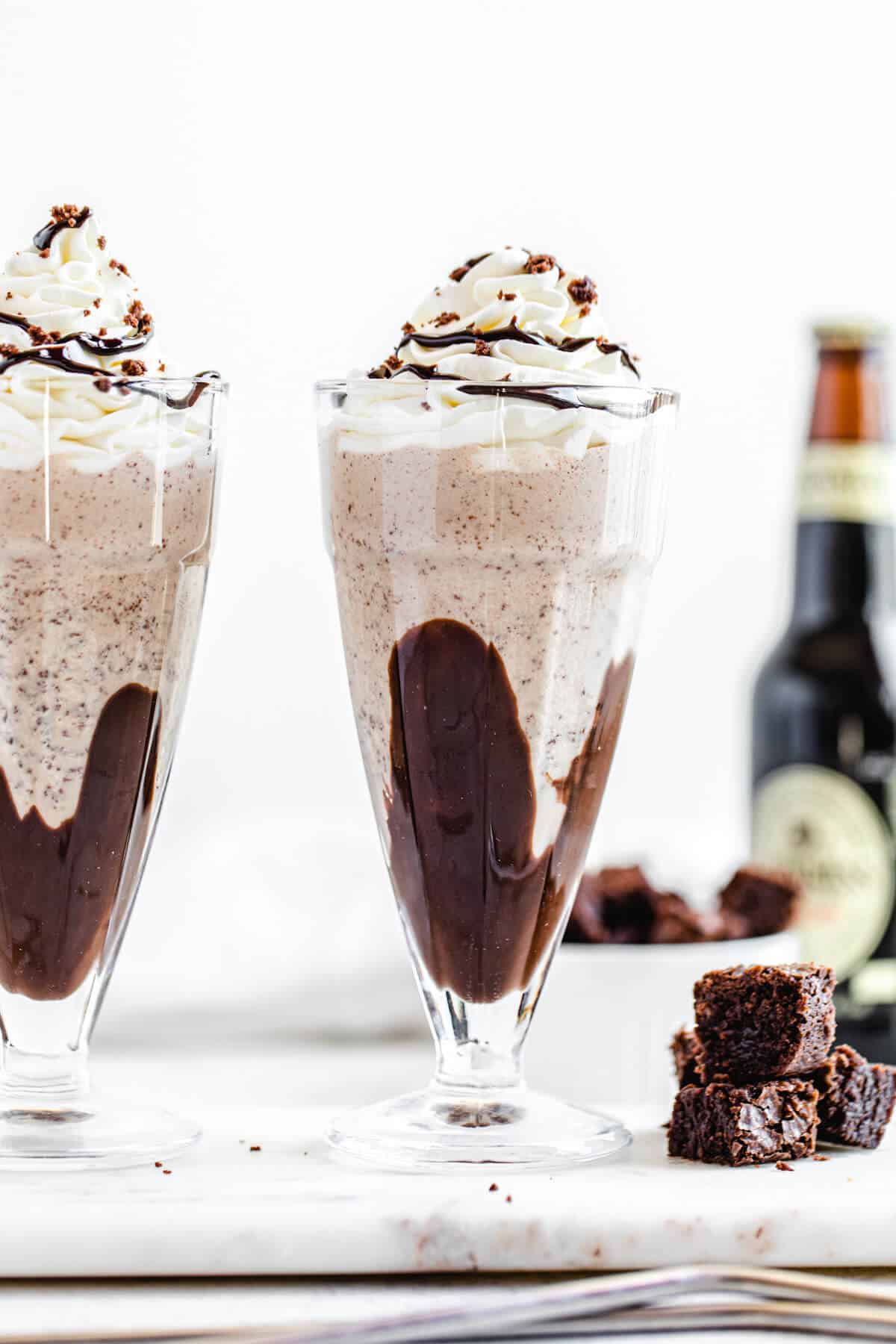 two milkshakes on a rectangle marble board with three brownie chunks beside them and a bottle of beer in the background