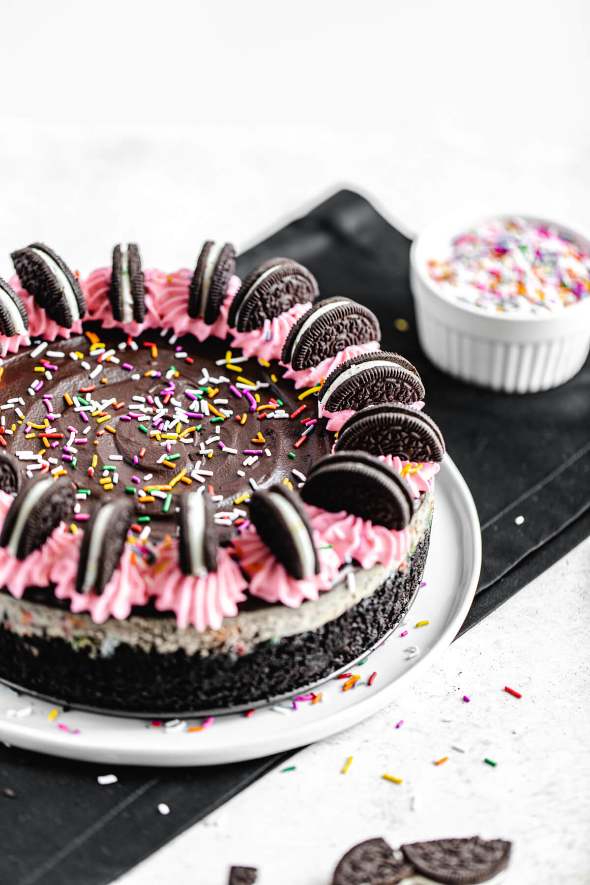 cheesecake on a white plate that is on a black silk napkin with a bowl of sprinkles in the background