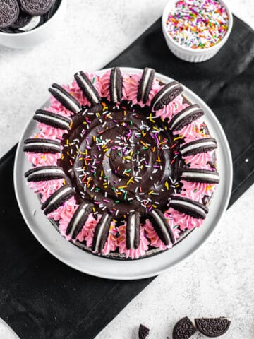 top view of cheesecake on a white plate that is on a rectangle black silk napkin with a bowl or Oreos and sprinkles on the side