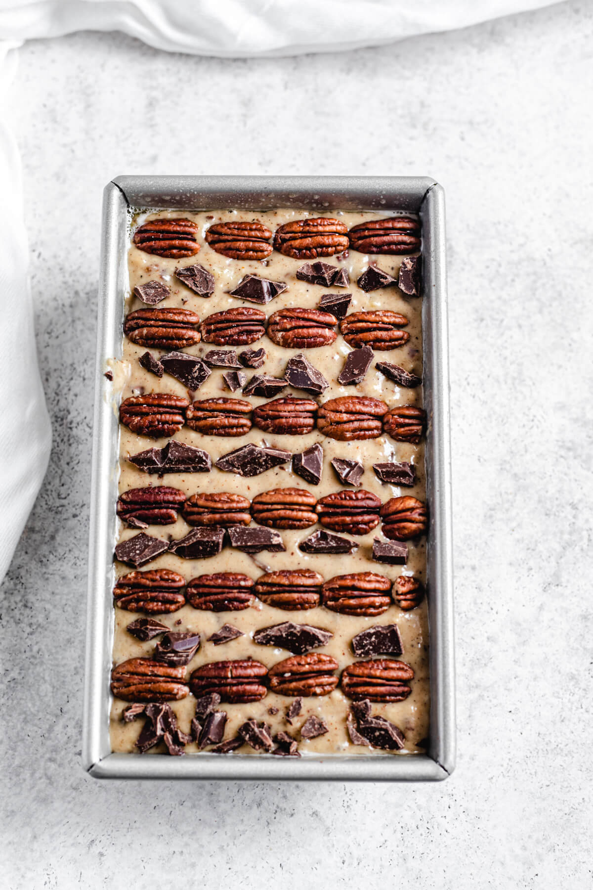 banana bread batter in a loaf pan with rows of pecans and chopped chocolate lined on top