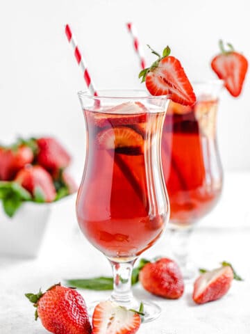 one glass of strawberry flavoured iced tea in the foreground and another glass in the background with fresh strawberries in front of the glasses and in a square white bowl in the background