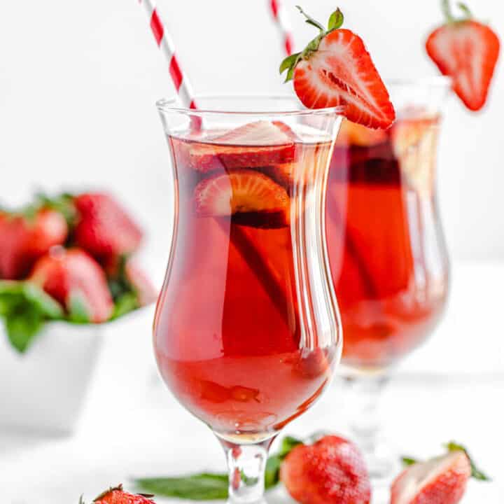 one glass of strawberry flavoured iced tea in the foreground and another glass in the background with fresh strawberries in front of the glasses and in a square white bowl in the background