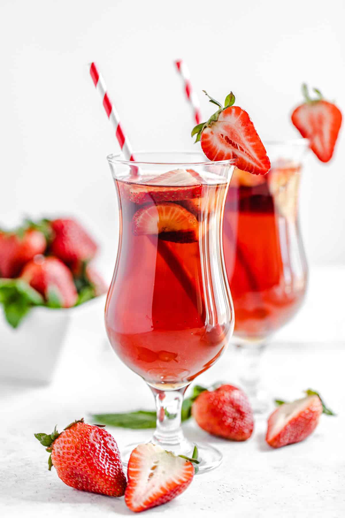 one glass of strawberry flavoured iced tea in the foreground and another glass in the background with fresh strawberries in front of the glasses and in a square white bowl in the background