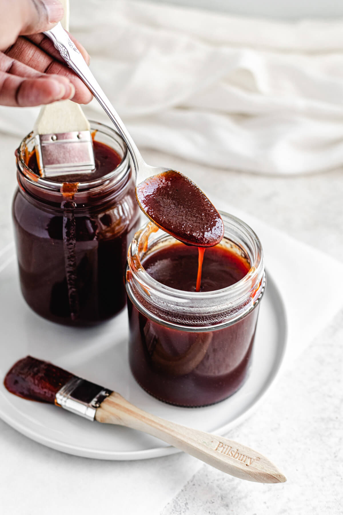 two jars of barbecue sauce on a white plate with a spoonful of sauce over one jar and a pastry brush on the side