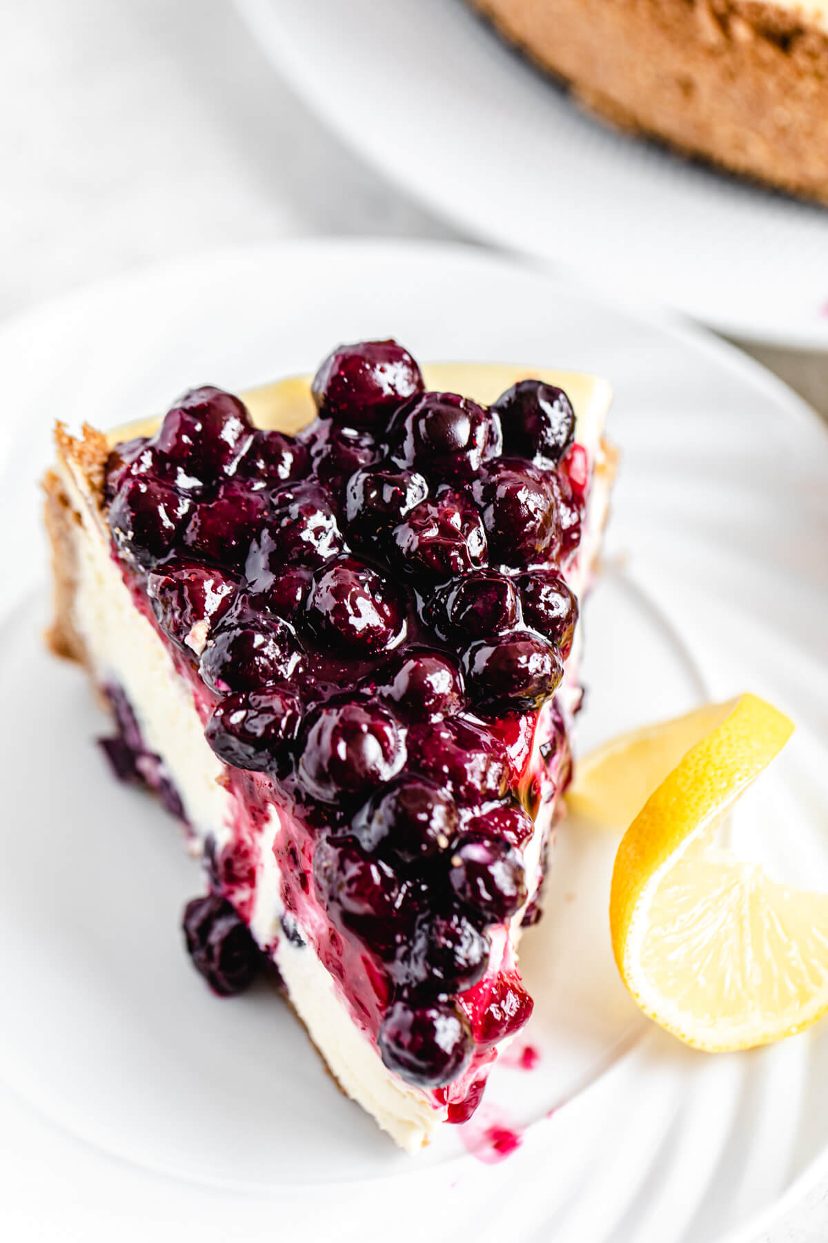 cheesecake slice topped with blueberry sauce on a white plate with lemon twist beside it