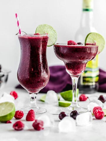 two glasses of daiquiris with ice cubes, cherries, raspberries and limes around it