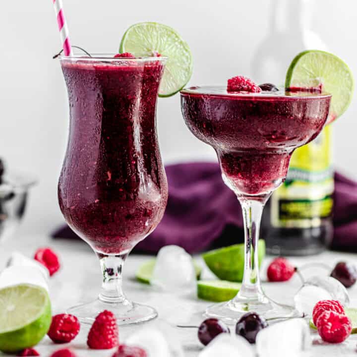 two glasses of daiquiris with ice cubes, cherries, raspberries and limes around it
