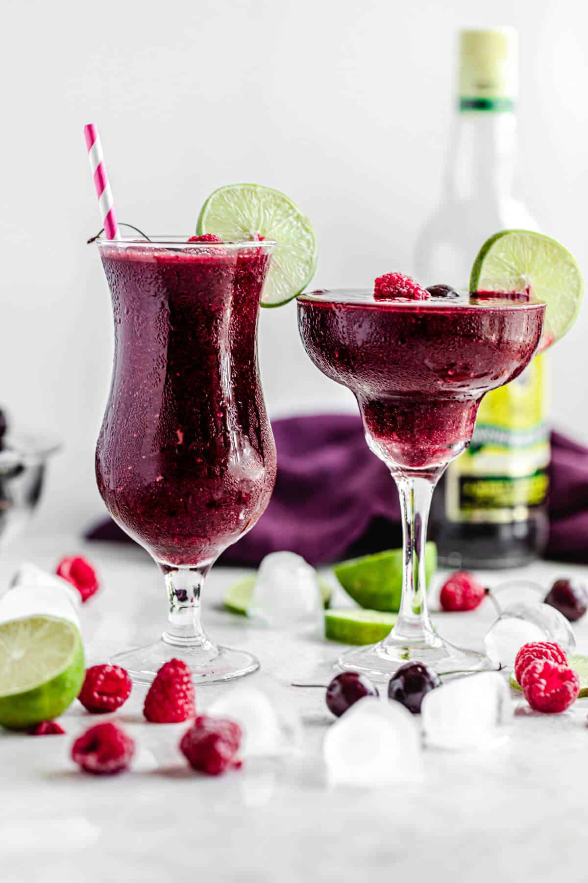 two glasses of daiquiris with ice cubes, cherries, raspberries and limes around it