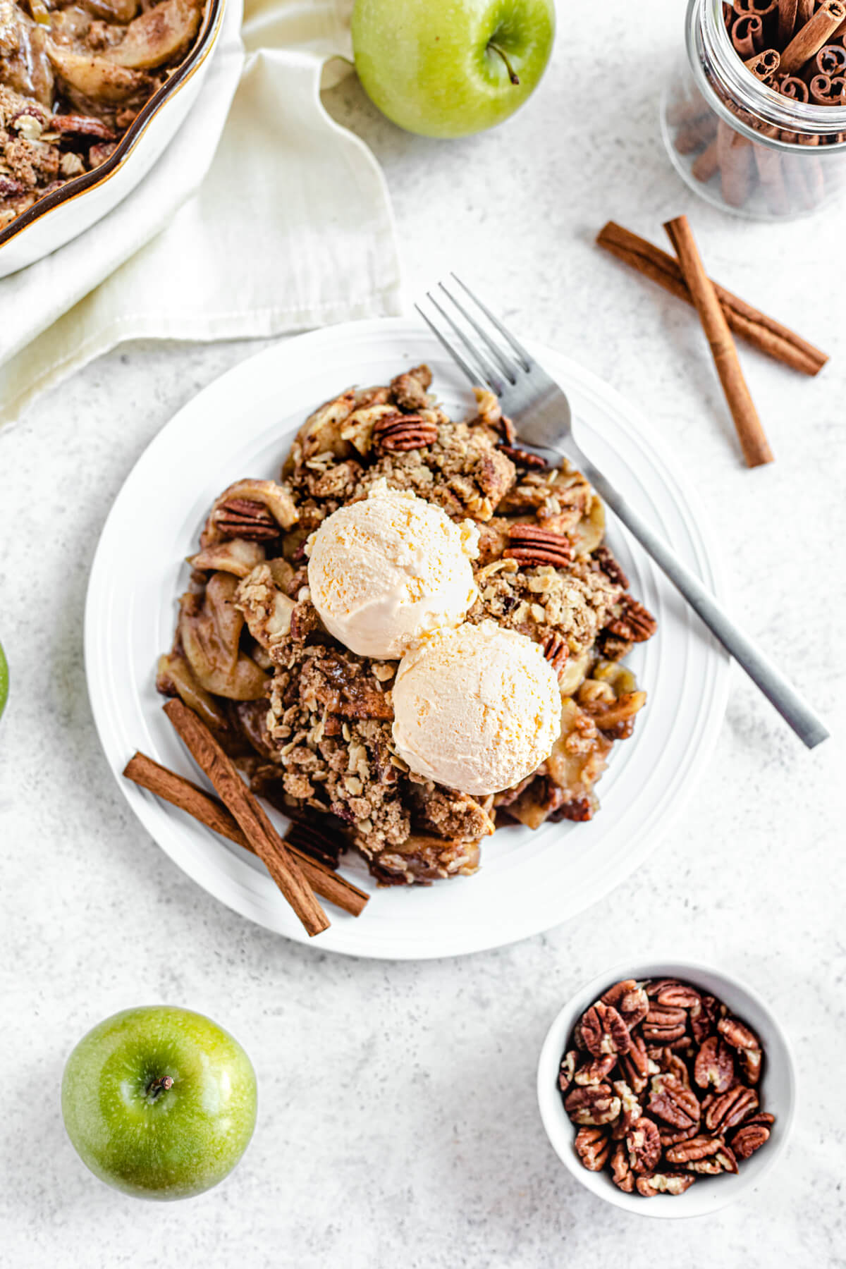 apple crisp topped with two scoops of ice cream in a white plate with two cinnamon sticks and a fork on the edges of the plate