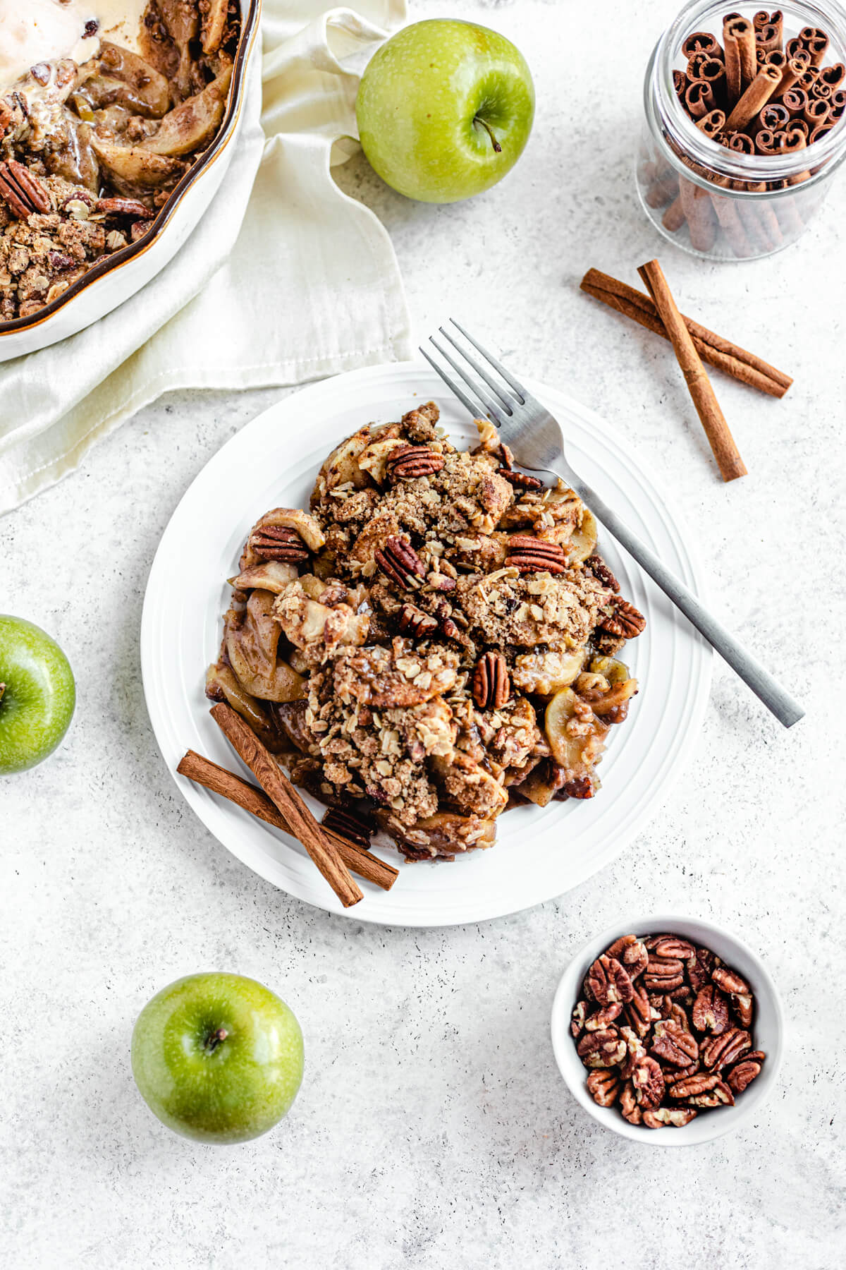 plain apple crisp in a white plate with two cinnamon sticks and a fork on the edges of the plate