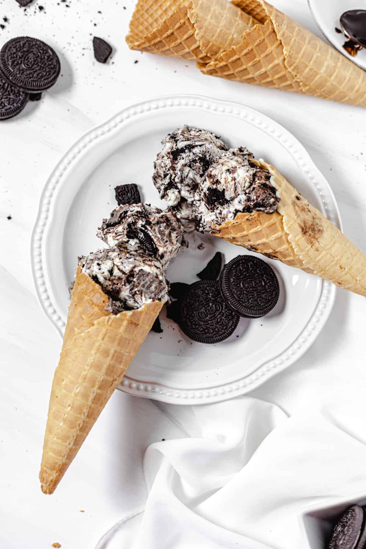 two cones filled with ice cream on a white plate with two Oreos on it and two waffle cones on the top right